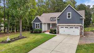 A gray two-story house with white trim and a double garage sits on a well-maintained lawn with shrubs, a concrete driveway, and surrounding trees. The sky is partly cloudy.
