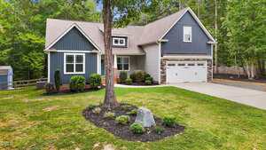 A gray and blue two-story house with a gabled roof, white trim, and a double garage sits behind a manicured lawn, mulch landscaping, and a tree in the front yard, bordered by woods in the background.