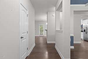 A hallway with white walls and dark wood flooring leads to a door with glass panes and a view outside. Openings in the wall reveal a kitchen with stainless steel appliances on the right.