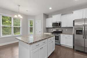 Modern kitchen with white cabinets, stainless steel appliances, marble countertop island, and wood flooring. Large windows and a hanging light fixture provide natural and artificial light.