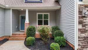 Front entryway of a house with gray siding, brick accents, a blue front door, and trimmed shrubs along a concrete walkway. A window is centered above the bushes.