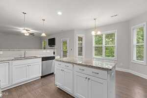 A modern kitchen with white cabinets, granite countertops, stainless steel appliances, an island in the center, wood flooring, pendant lights, and large windows with views of greenery outside.