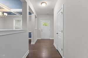 A hallway with light-colored walls, a white front door with glass panels, hardwood floors, a ceiling light, and two closed white doors on the right. An open dining area with a chandelier is visible on the left.