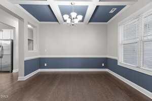 A dining room with blue and white coffered ceiling, blue and white walls, dark wood floors, a modern chandelier, a large window with blinds, and an opening leading to a kitchen with stainless steel appliances.