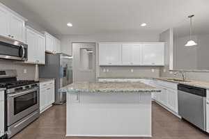 A modern kitchen with white cabinets, stainless steel appliances, a center island with a granite countertop, and light brown wood flooring. The space is well-lit with recessed lighting and a pendant light over the sink.
