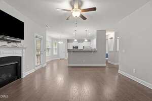 Open-concept living area with hardwood floors, ceiling fans, a fireplace with a TV above it, and a kitchen with white cabinets and pendant lighting in the background. Glass doors open to the outside.