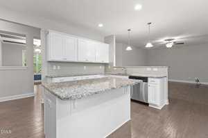 Modern kitchen with white cabinets, granite countertops, stainless steel appliances, and pendant lighting, open to a living area with wood flooring and a ceiling fan. Neutral wall colors and recessed lighting are visible.