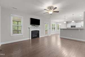 A spacious open-concept living area with hardwood floors, a ceiling fan, a wall-mounted TV above a fireplace, large windows, and a view of a modern kitchen with white cabinets and stainless steel appliances.