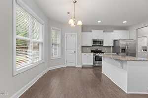 Bright kitchen with white cabinets, stainless steel appliances, granite countertops, and a central island. There is a pantry door and large windows with blinds overlooking a green outdoor area. The floor is dark wood.