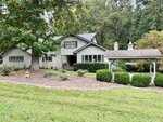 A gray two-story house with stone accents sits on a sloped lawn at 100 Maluli Drive, Oxford, surrounded by trees and bushes. A small white gazebo-style structure with a weathervane is on the right side of the property.