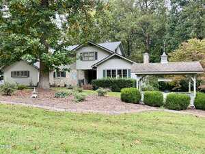 A gray two-story house with stone accents sits on a sloped lawn at 100 Maluli Drive, Oxford, surrounded by trees and bushes. A small white gazebo-style structure with a weathervane is on the right side of the property.