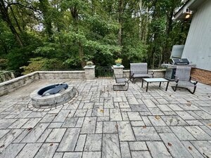 Stone patio with a built-in fire pit, outdoor furniture including chairs and a grill, bordered by a low stone wall and surrounded by dense trees in the background.