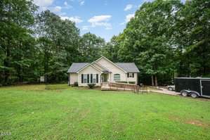 A single-story beige house with white trim sits in a large grassy yard at 2488 Patton Lane, Franklinton. Surrounded by tall trees, it features a wooden ramp to the front door and a black trailer parked on the right side.