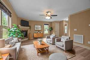 A living room with beige walls, light wood flooring, and a ceiling fan. The furniture includes a gray sofa, two gray armchairs, a wooden coffee table, and a sideboard, with plants and framed art for decor.