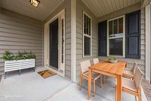 A covered porch with beige siding features a black front door, a welcome mat, a white planter with greenery, and a wooden dining table with four woven chairs and a small potted plant centerpiece.