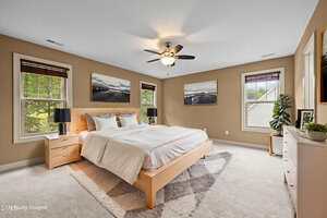 A spacious bedroom with beige walls, a large bed with white bedding, two nightstands with lamps, three landscape photos on the wall, carpeted floor, ceiling fan, and multiple windows letting in natural light.