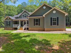 A charming single-story house at 5300 Langdon Drive, Oxford, with gray siding, white trim, black shutters, and a brick foundation. Enjoy the covered front porch surrounded by trees and lush green grass.