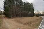 A rural intersection with a dirt road leading into a wooded area on the left and a paved road curving right; there are trees, utility poles, and a small street sign visible at the corner.