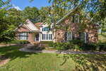 A two-story suburban house with red brick and white siding sits at 3538 Saddle Ridge Road, surrounded by green grass, manicured bushes, and trees under a blue sky with scattered clouds.