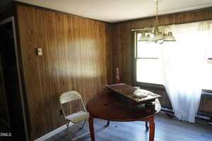 A small room with wood-paneled walls, a round wooden table with items on top, a folded white metal chair, and a window with sheer curtains letting in sunlight. A ceiling light fixture hangs above the table.