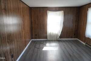 An empty room with wood-paneled walls, gray laminate flooring, a window covered by a sheer curtain letting in sunlight, and loose black cables on the floor near the wall.