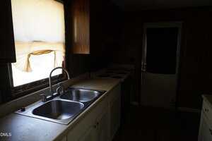 A dimly lit kitchen with a double sink, white cabinets, an electric stove, and a countertop. Sunlight comes through a window with a torn curtain. A closed door is visible in the background.