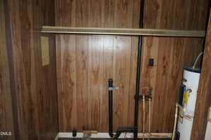 A small utility room with wood-paneled walls, exposed plumbing pipes, a water heater on the right, and a horizontal wooden board mounted above the pipes. A paper is attached to the left wall.