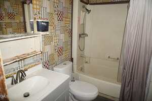 A small bathroom with patterned wallpaper, a white sink with a faucet, a toilet, and a cream-colored bathtub with a showerhead and grab bar. The wall shelf above the sink has visible rust and wear.