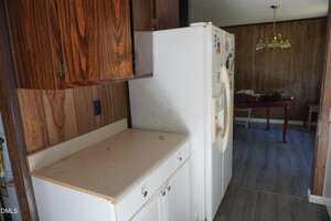 A kitchen with wood-paneled walls, stained countertop, and a white refrigerator with visible dirt. A dining area with a table and gold chandelier is partially visible in the background.