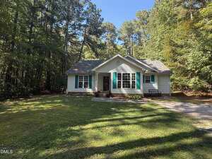 A small white house with teal shutters and a shingled roof sits in a grassy yard surrounded by tall trees. A curved path leads to the front door, with a bench and flowerbed near the entrance.