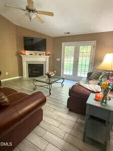 A living room with two brown leather sofas, a glass coffee table, a fireplace with a TV above it, a side table with a lamp and flowers, and glass double doors leading outside. The floor is light wood-style tile.