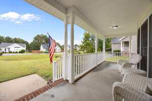A covered front porch with white railings and wicker chairs overlooks a green lawn and an American flag. Neighboring houses are visible across a quiet residential street under a partly cloudy sky.
