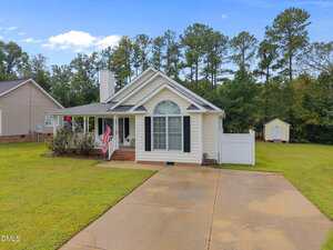 Single-story house with a large arched window, covered front porch, and American flag. There is a concrete driveway, a white fence, and a backyard shed, with trees in the background on a cloudy day.