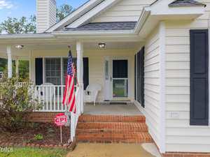 A white house with black shutters features a covered front porch, two white chairs, a brick step entry, an American flag, a red CPI security sign in the yard, and some shrubs near the porch.