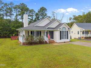 Single-story white house with a gray roof, front porch, and American flag. The house has several windows and is surrounded by grass and trees. A concrete driveway is visible in the foreground.