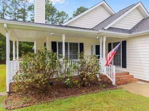 A white house with black shutters features a covered front porch with white railings, brick steps, an American flag, and bushes in front. The lawn is green and trees are visible in the background.