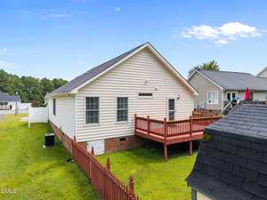 A single-story house with white siding and a dark roof features a wooden deck and fenced backyard. The yard is grassy, and neighboring houses are visible, with a blue sky and scattered clouds above.