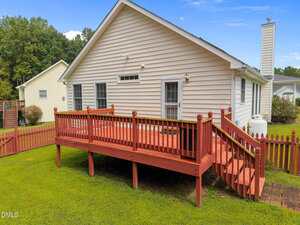 A beige single-story house with white trim features a wooden raised deck with stairs and a red railing, surrounded by a red picket fence. The yard has green grass, and trees are visible in the background.