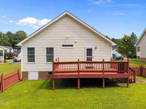 A single-story house with beige siding and brick foundation, featuring a raised wooden deck with red railing, set in a grassy backyard and bordered by a matching red fence.
