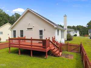 Single-story house with light-colored siding, a red wooden deck with steps leading to a fenced backyard, green lawn, and trees in the background under a clear blue sky.