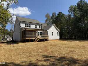 A two-story white house with a screened-in porch and wooden deck sits on a newly cleared lot with straw covering the ground, bordered by tall trees under a blue sky.