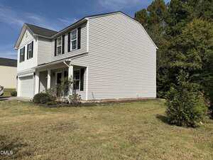 A two-story beige house with white trim, black shutters, and a covered front porch. The house has a two-car garage and sits on a grassy lawn with a few shrubs and trees around the property.