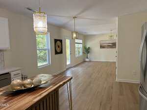 A bright, open-concept interior with light wood flooring, cream walls, pendant lights, large windows, a door to the outside, and minimal decor including a plant and a decorative bowl on a wooden island.