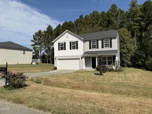 Two-story white house with black shutters and a gray roof, attached two-car garage, front porch, and a well-kept lawn with some bushes and trees, set against a backdrop of tall pine trees under a blue sky.