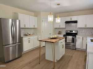 Modern kitchen with white cabinets, stainless steel appliances, a wooden island with gold legs, light wood flooring, and two pendant lights hanging above the island. Marble-patterned backsplash and countertop are visible.