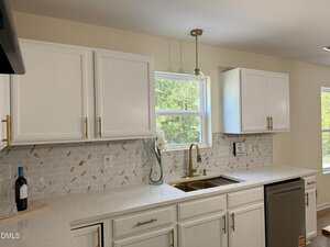 A modern kitchen with white cabinets, gold hardware, a white marble countertop, geometric tile backsplash, a stainless steel dishwasher, gold faucet, wine bottle, vase with white flowers, and a window above the sink.