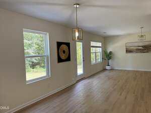 A bright room with light wood flooring, cream-colored walls, large windows, a glass door, two modern pendant lights, wall art, and a potted plant. Trees are visible outside through the windows.