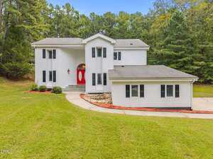 A two-story white house with black shutters and a red front door at 141 Southerland Place in Henderson, surrounded by grass and trees. The curved driveway leads to landscaped areas with rocks and mulch near the entrance.