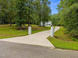 A long concrete driveway with two white pillars at the entrance leads through a grassy, wooded area to a white two-story house in the background. The sky is clear and blue.