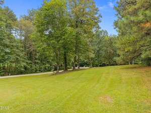 A grassy lawn slopes gently toward a paved road, bordered by tall green trees under a bright blue sky with scattered clouds.